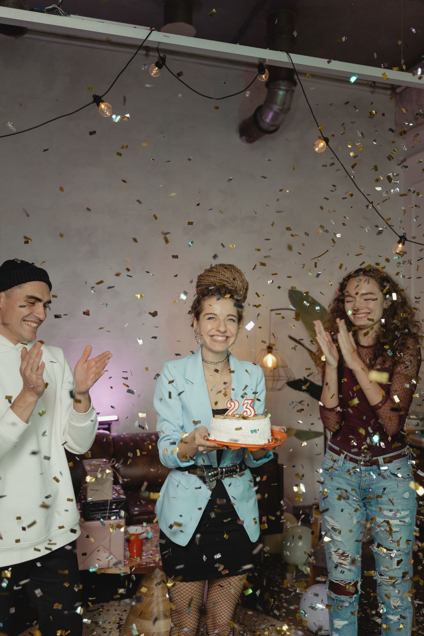 Group of young adults celebrating a 25th birthday with cake and confetti indoors.